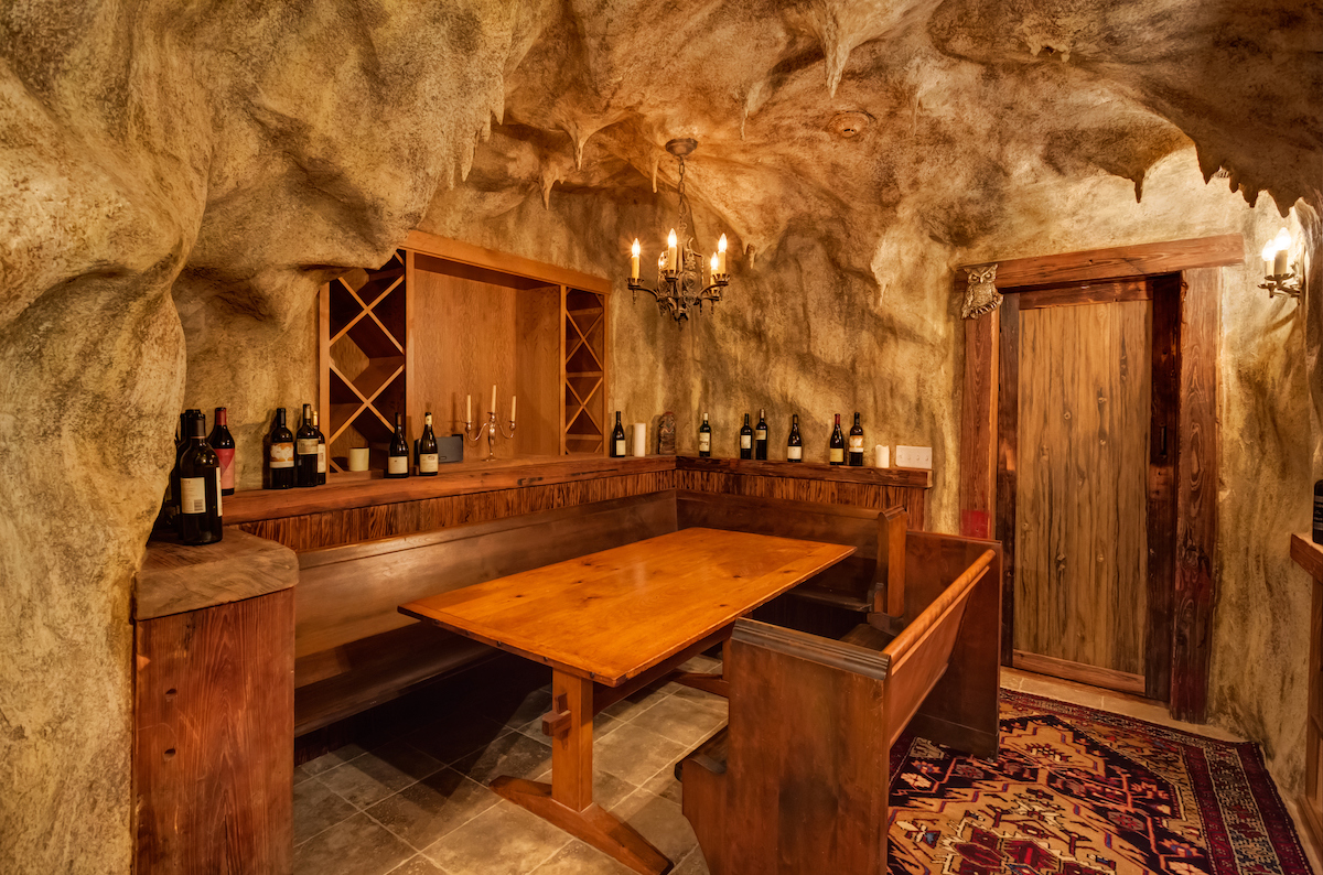 A wine cellar with a wooden door, stone tile flooring, a rug, and a wooden table and bench is located within cave walls.