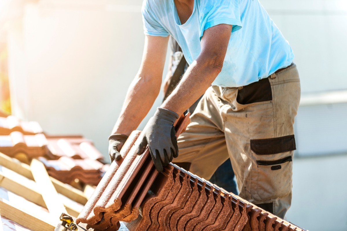 A worker in gloves and a blue shirt hauls heavy roofing materials.