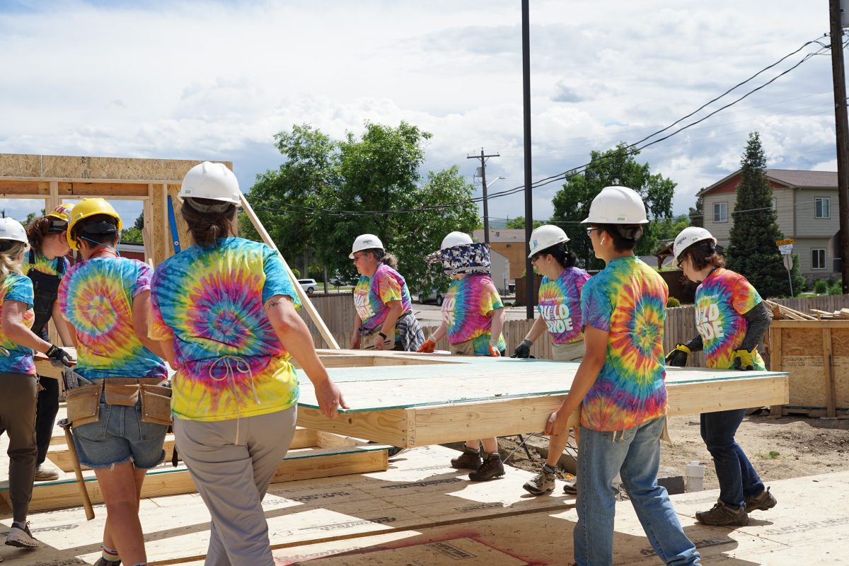 A group of Pride Month Metro Denver volunteers carry a completed wall frame across a job site.