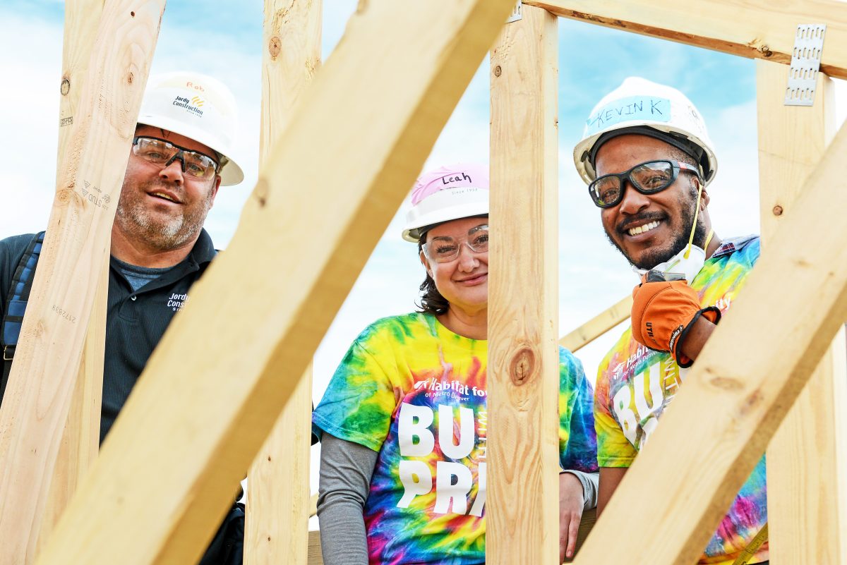 A trio of Pride Build Metro Denver volunteers pose behind the framed wall of an in-construction house.