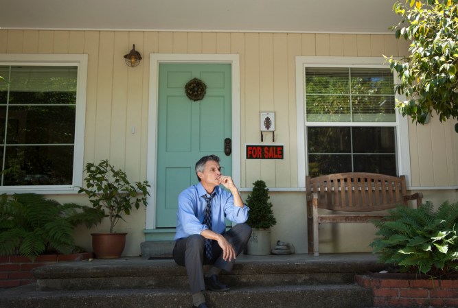 A man sits in front of a home with a green door.