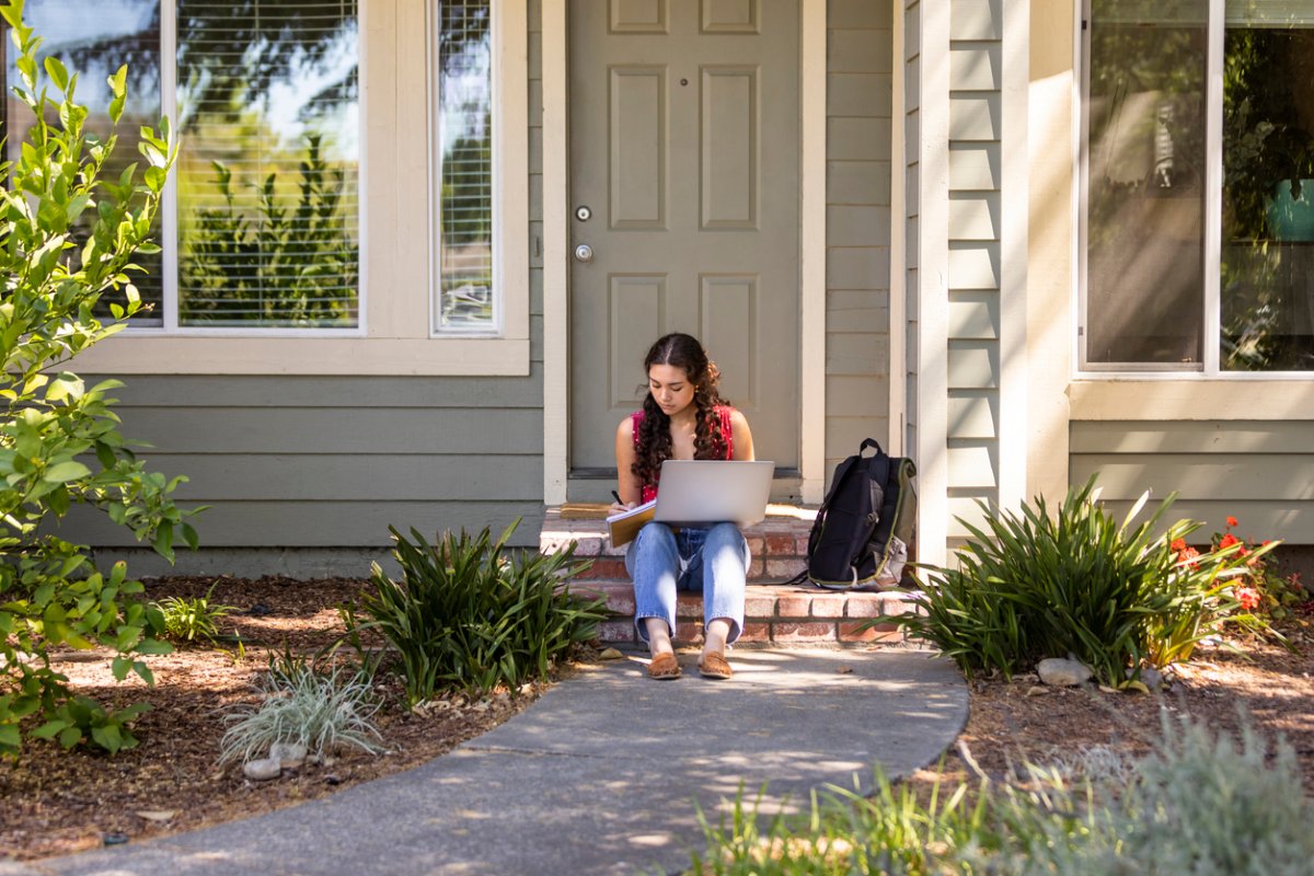 A woman sits in front of a grey house while on a laptop, writing in a notebook. 