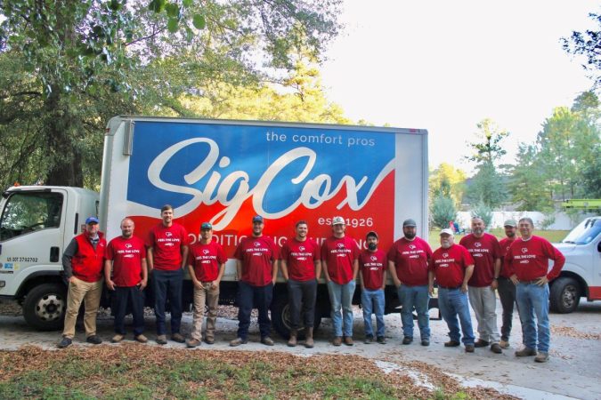 The Sig Cox team of thirteen men wearing matching red "Feel the Love" Lennox shirts are standing in front a Sig Cox truck.