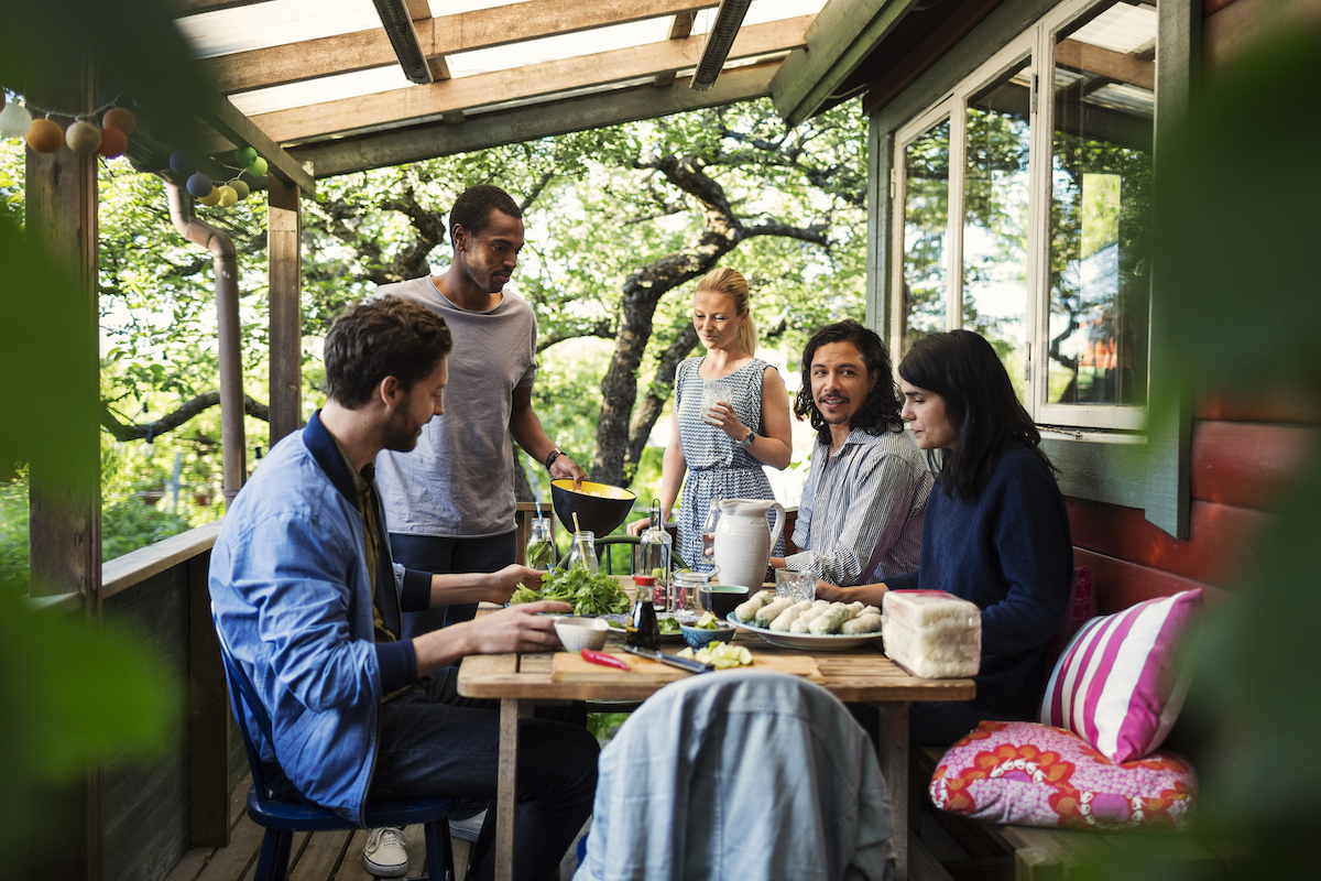 A group of friends gather on the porch of a home, with a shaded roof overhead.
