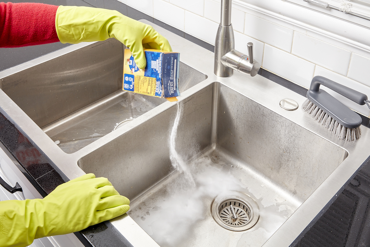 Woman wearing rubber gloves sprinkles baking soda from a box on a stainless steel sink.