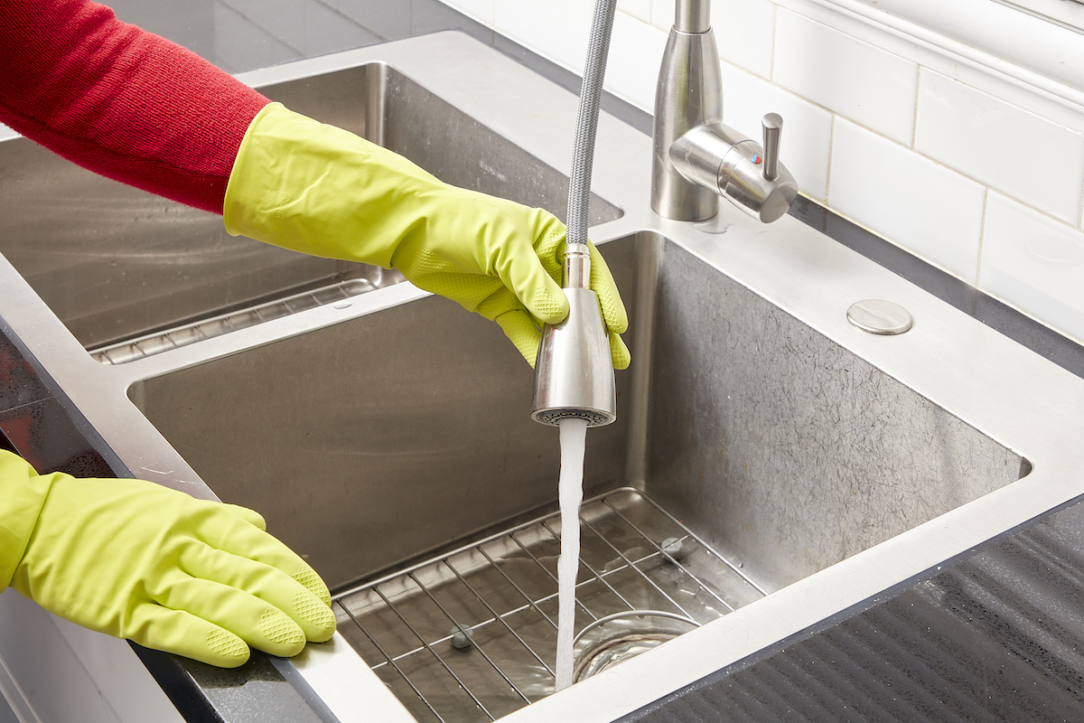 Woman wearing yellow rubber gloves rinses a stainless steel sink with the sink faucet.