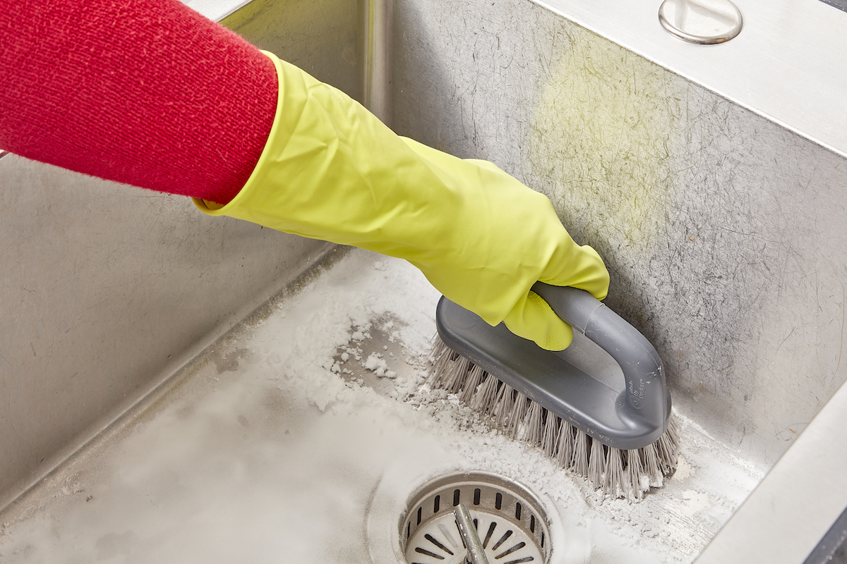 Woman wearing rubber gloves uses a scrub brush to scrub baking soda in a stainless sink.