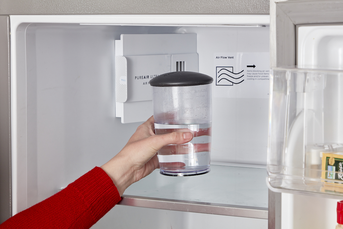 Woman wearing red sweater puts a glass container with clear liquid into the refrigerator.