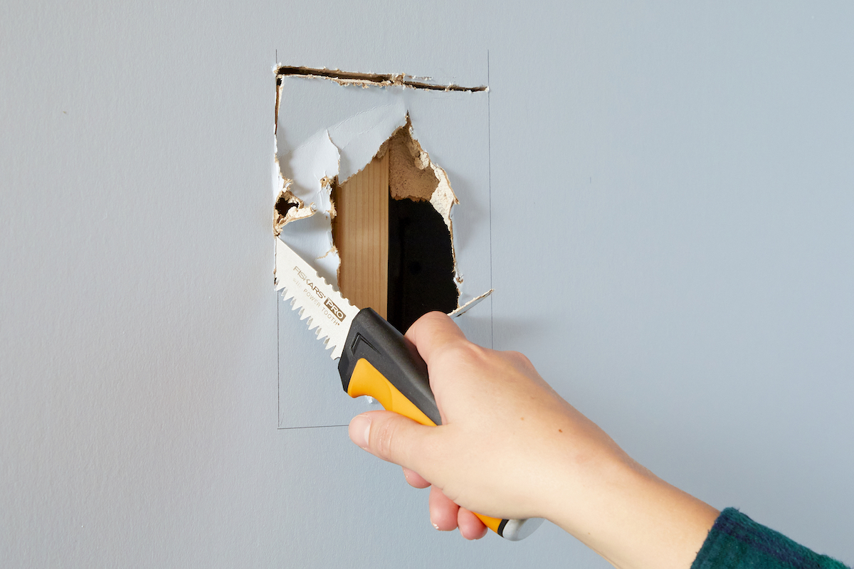 Woman uses drywall saw to cut a square around large hole in drywall.