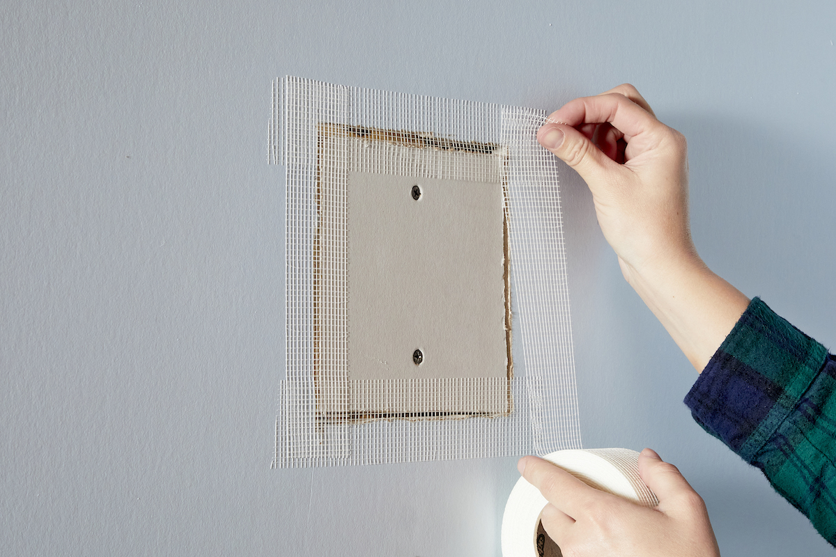 Woman tapes edges of a drywall patch with drywall mesh tape.