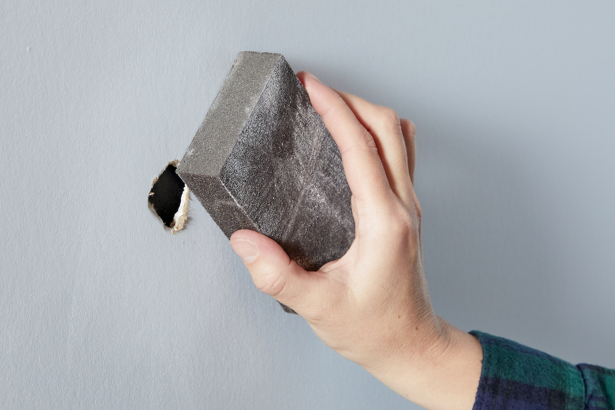 Woman uses a sanding block to sand a medium-sized hole in drywall.