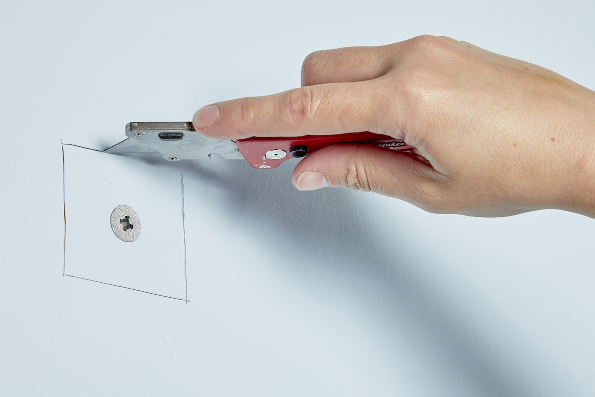Woman uses a utility knife to score a square area around a drywall anchor.