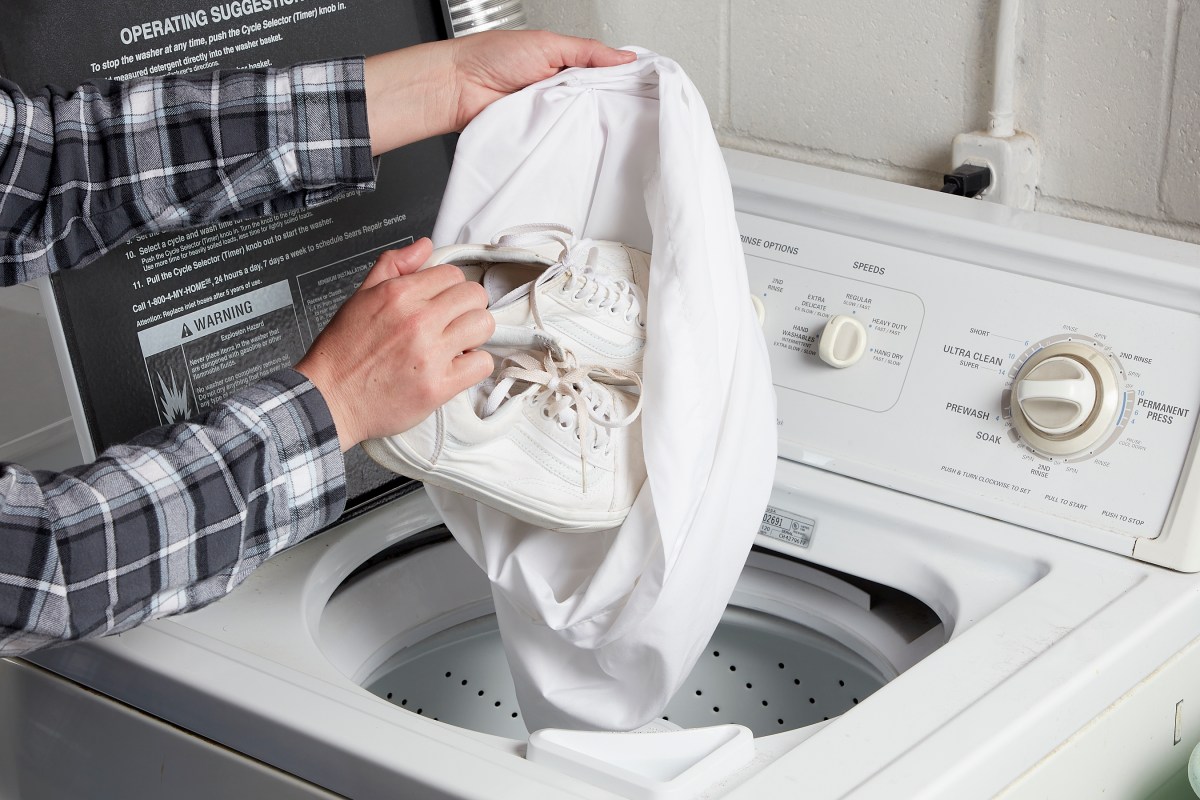 Woman places a pair of white canvas shoes in a white pillowcase, and prepares to toss both into a top-load washer.
