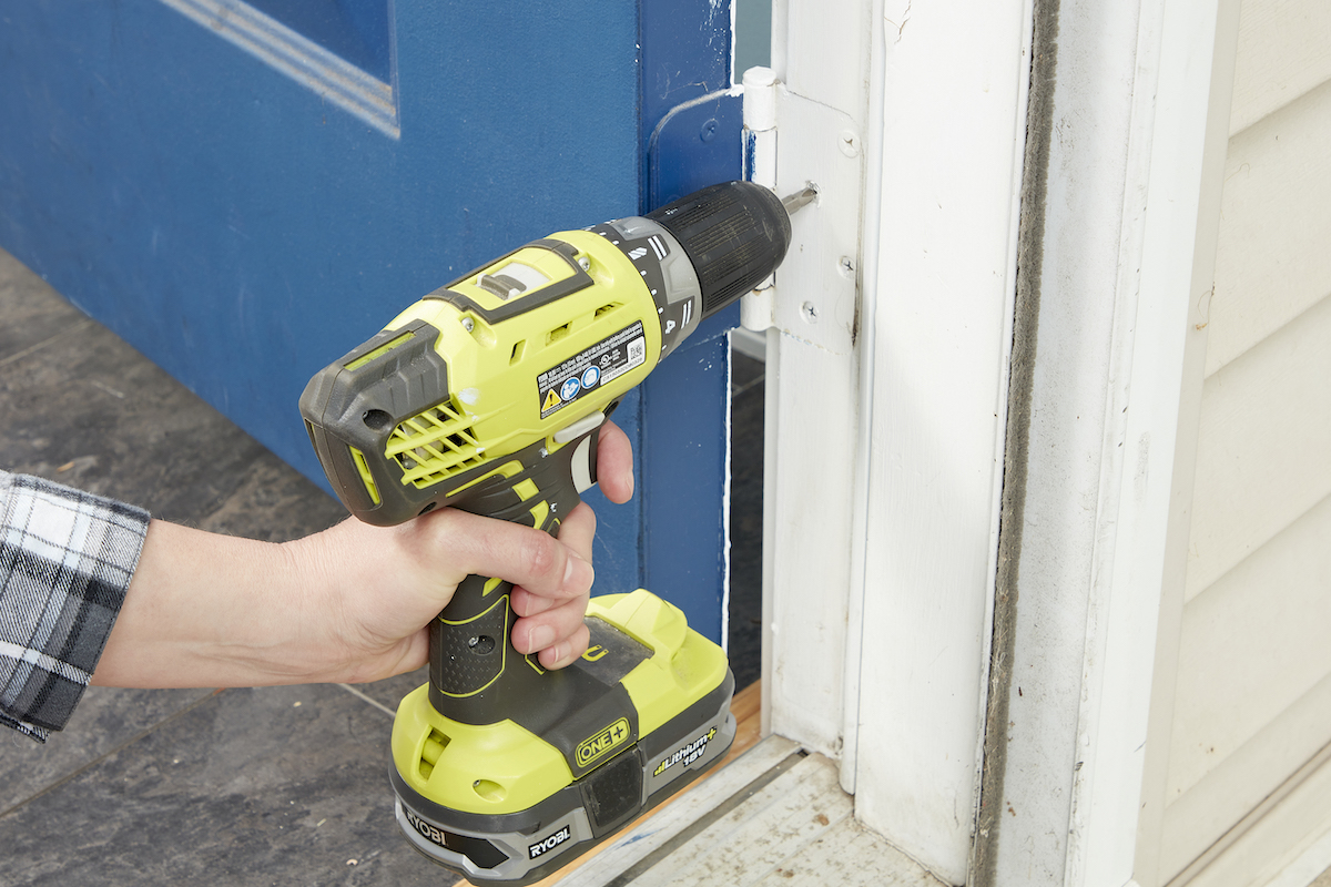 Woman tightens bottom hinges on an exterior door with an electric screwdriver.