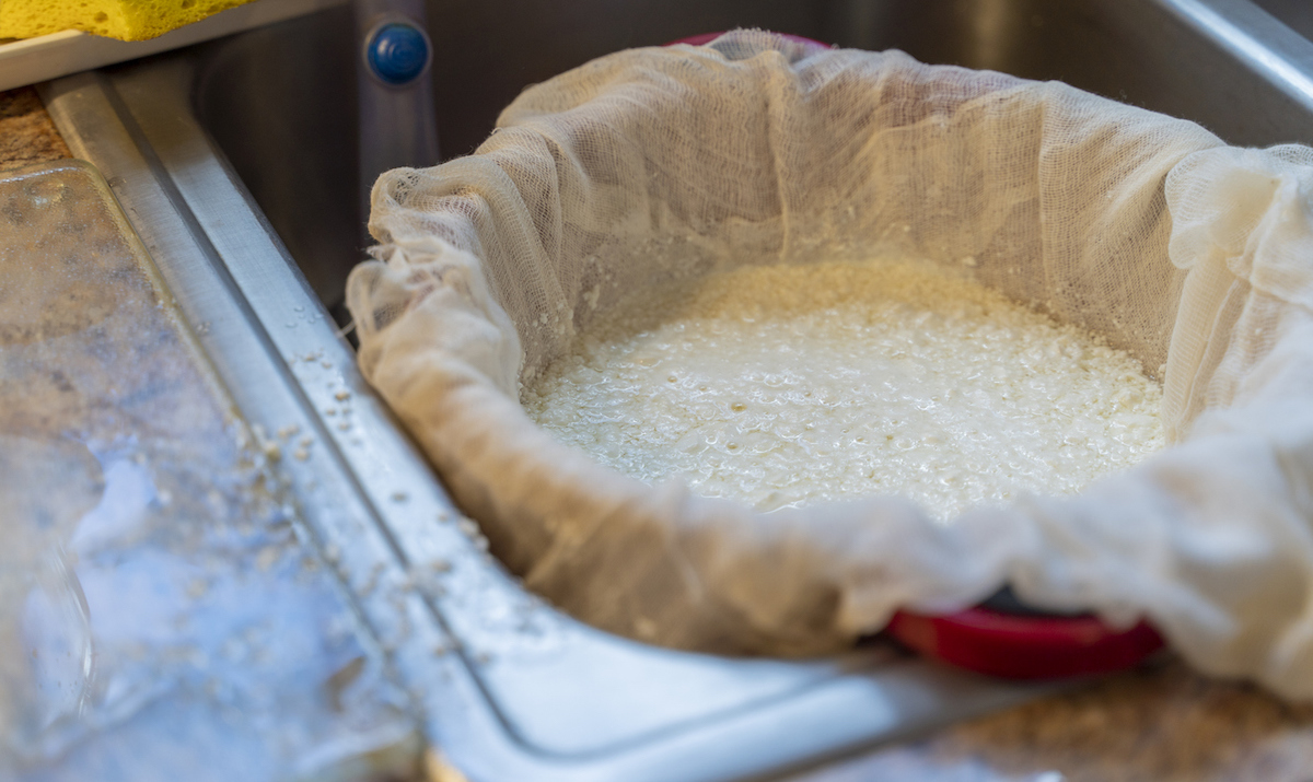 Curdled milk is inside of a cheesecloth-lined red plastic colander that is placed in a kitchen sink.