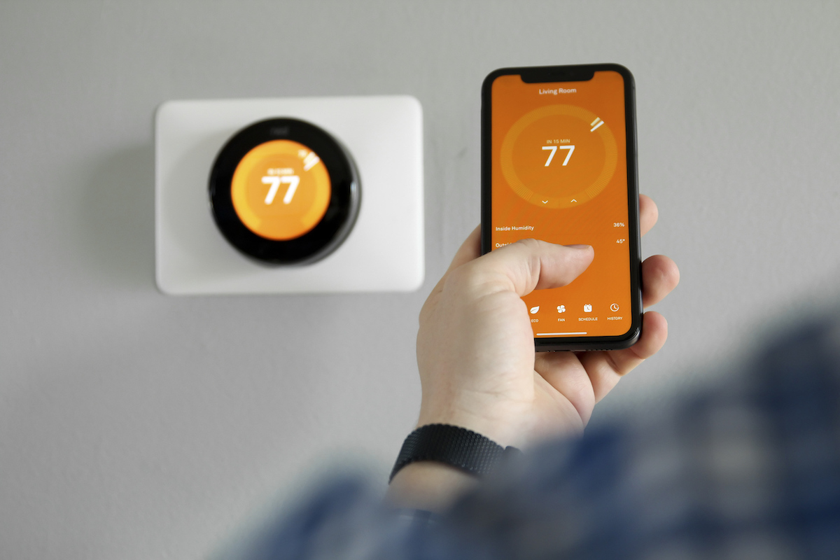 A person is using an orange thermostat app on their smart phone to change the smart thermostat to 77 degrees Fahrenheit.