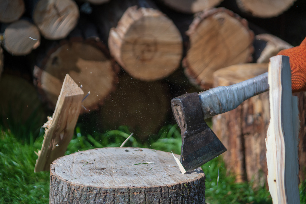 A person is splitting firewood with a splitting maul while wearing gloves and firewood is stacked in the background.