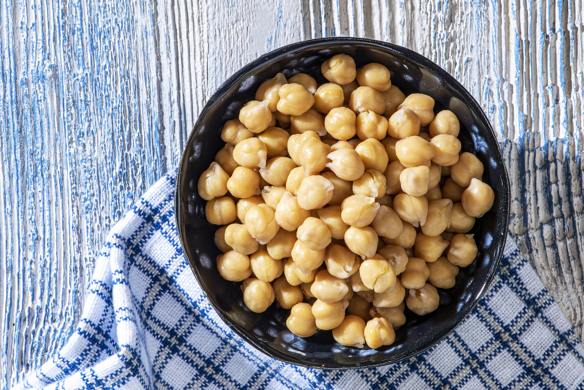Boiled chickpeas are in a dark bowl on a blue and white patterned towel on a white wooden surface.