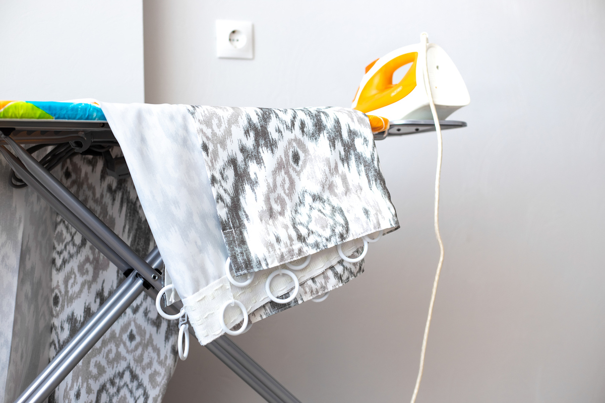 Grey and white patterned curtains and an orange iron are on top of an iron board.