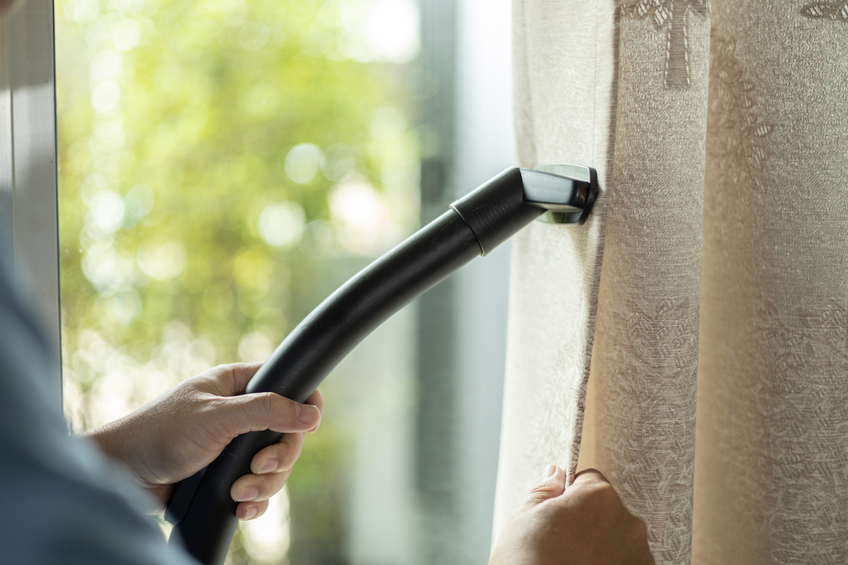 A person is using a vacuum cleaner attachment to clean hanging beige curtains at a window.