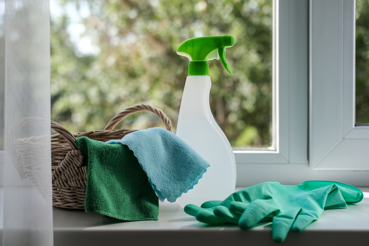 A rattan basket with clean rags inside, a spray bottle, and a pair of green rubber gloves are on a windowsill.