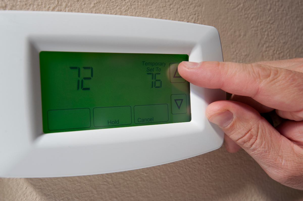 A person is increasing the temperature on a digital thermostat from 72 degrees Fahrenheit to 76 or higher.