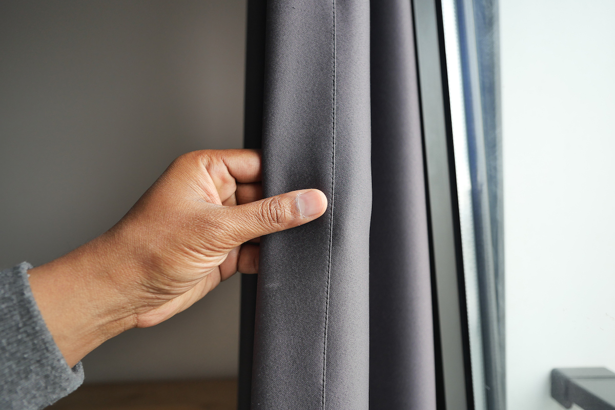 A person is holding a hanging grey curtain open in a window.