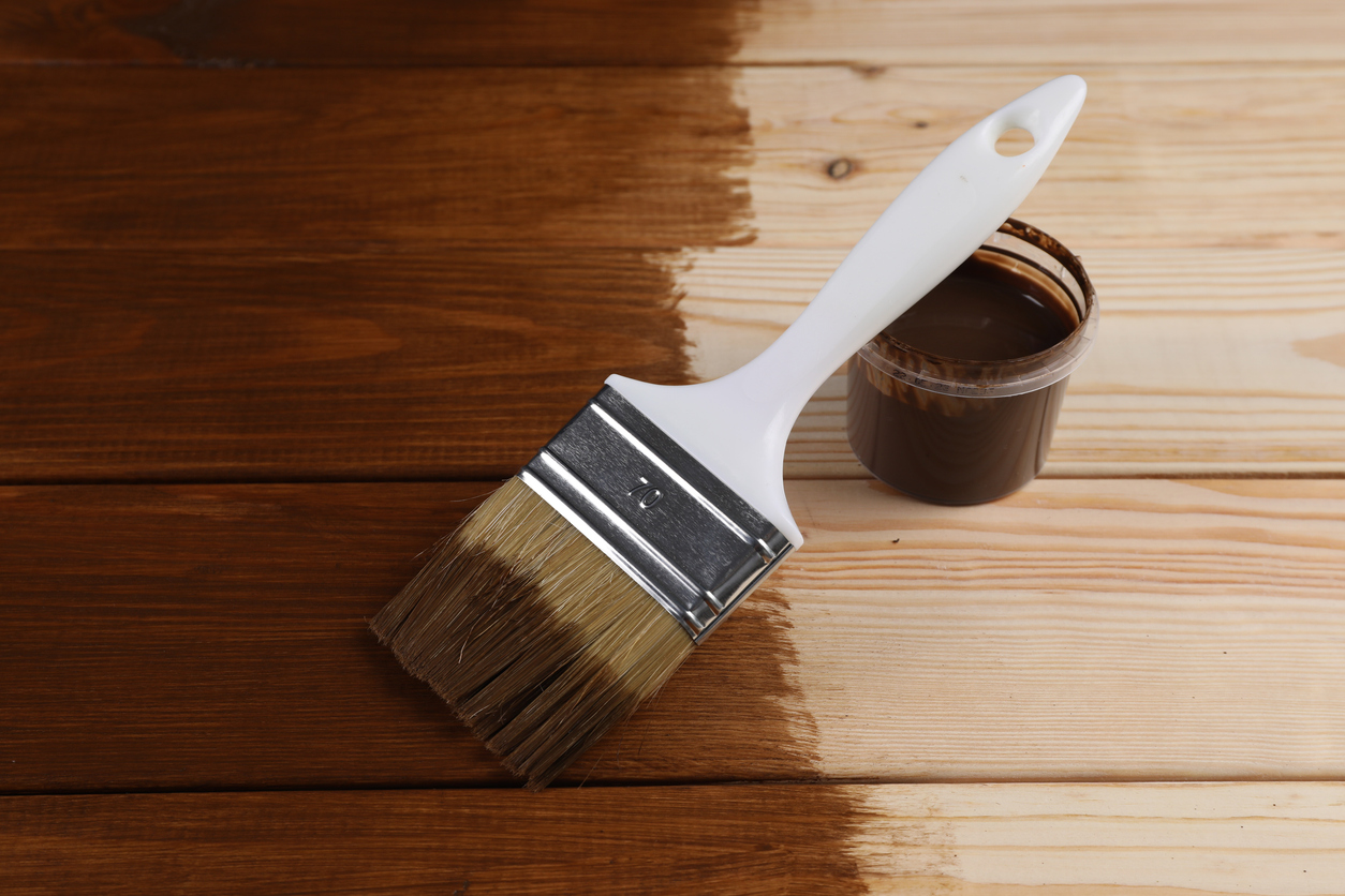 A wet paintbrush and a plastic container of dark-brown wood dye sit atop a half-dyed wooden tabletop.