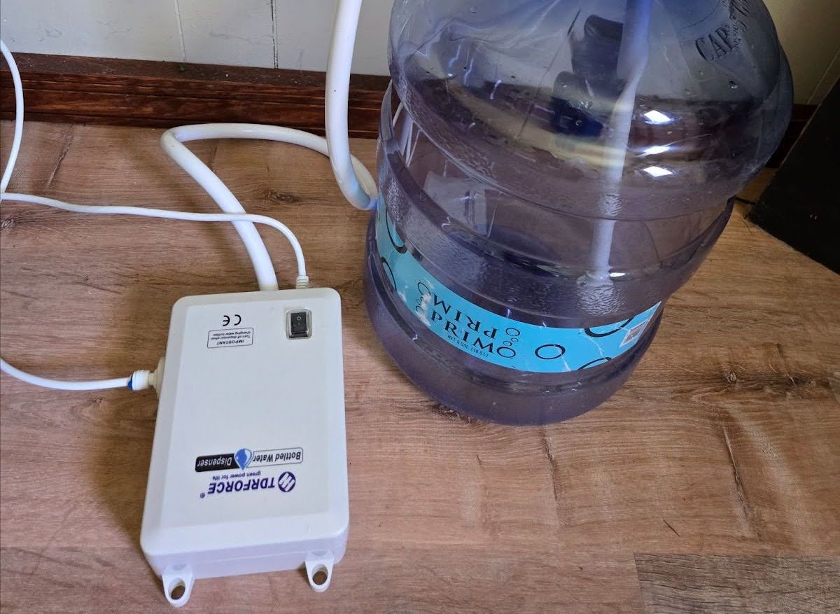 A bottled water pump system is sitting next to a 5-gallon water jug on a garage floor.