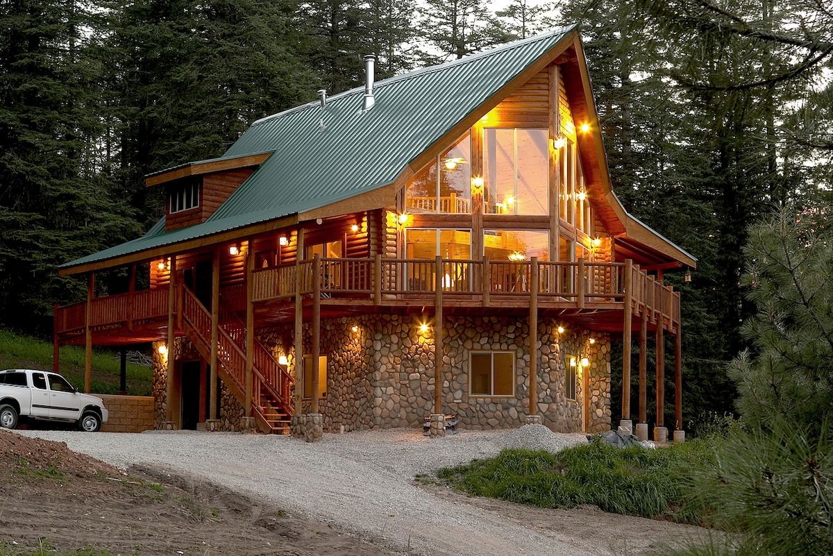 A log cabin lit up with lights in the evening with a green metal roof.