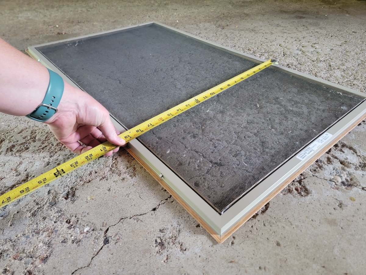 A person measuring a window screen to vent a portable AC unit in a casement window.