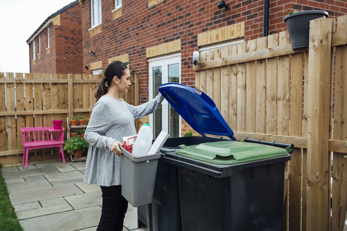 A person putting plastic and cardboard containers into a recycling bin in the backyard.