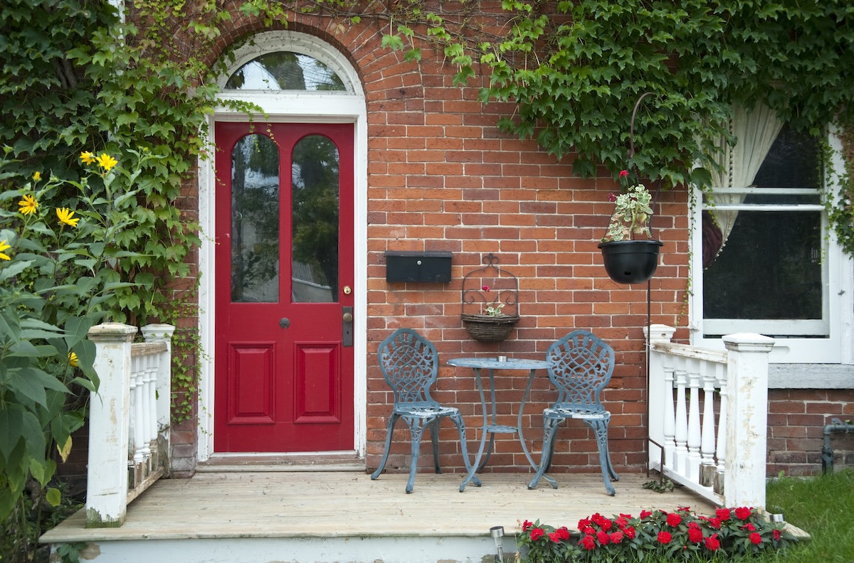 A brick house with a red front door and small front porch decorated with a table and chairs and plants.