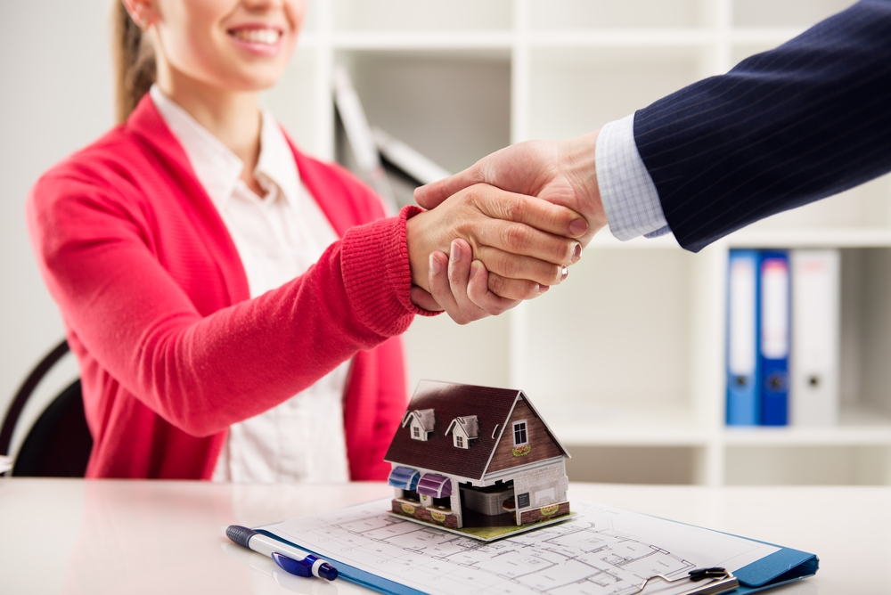 Two people shake hands over a small model house on a house plan on a clipboard. 