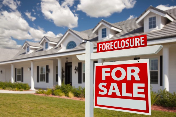 A red "Forelosure For Sale" sign is displayed in front of a house on a bright day.
