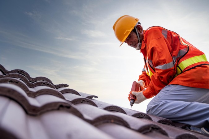 A roofer in an orange jacket and yellow hardhat is seen on a slate roof.