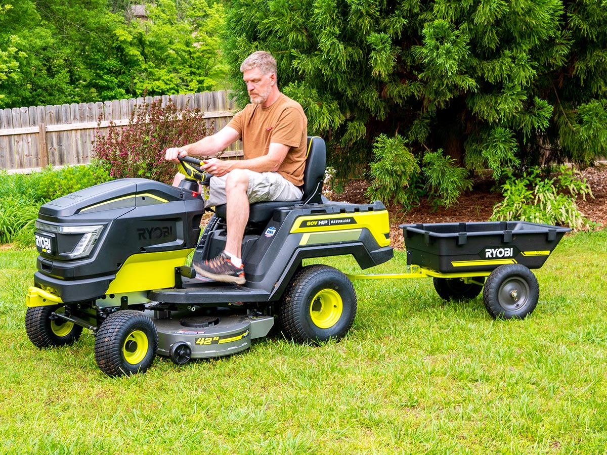 Person in brown t shirt driving Ryobi 80V HP Electric Lawn Tractor on grass in front of trees