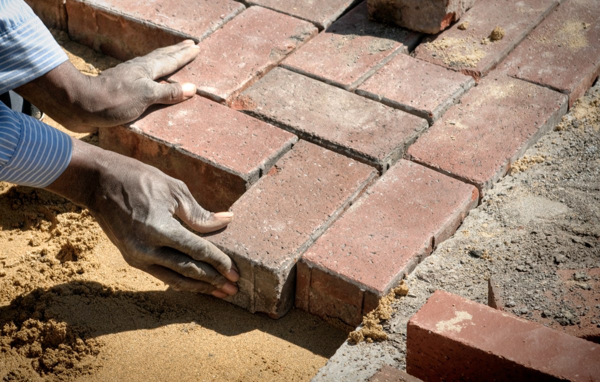 Black man's hands laying a red brick patio in a herringbone pattern.