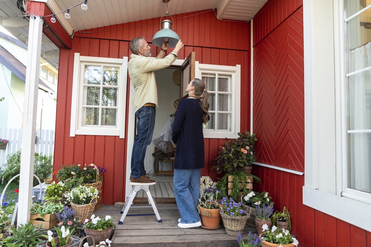 Man and woman changing lamp light bulb on porch of red home.
