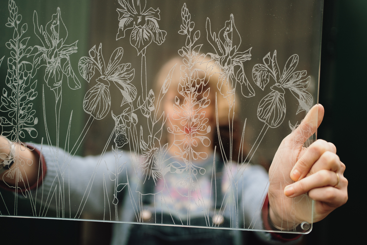 Shot looking through etched glass panel with intricate meadow design. Woman holding it up is out of focus on the other side.
