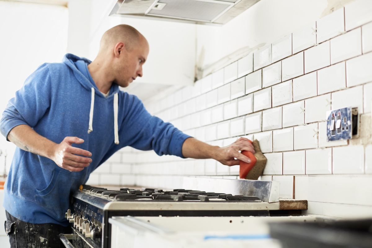 A man wearing a blue hoodie applies grout to kitchen backsplash.