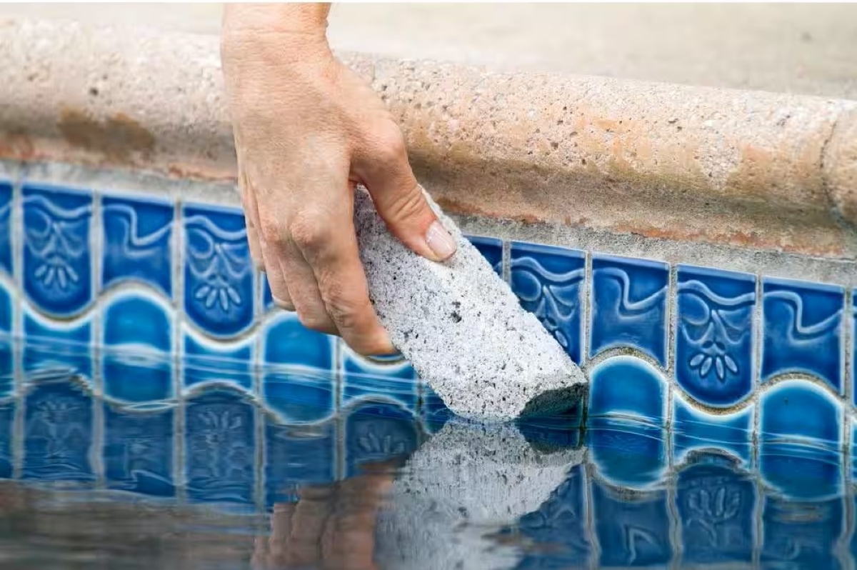 A hand is using a pumice stone to clean black algae from the pool tiles.