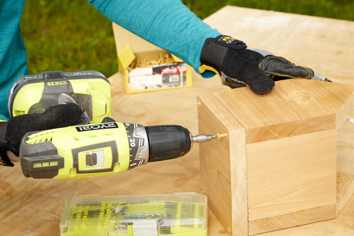 Woman uses a drill to screw the bottom of a birdhouse onto the sides.