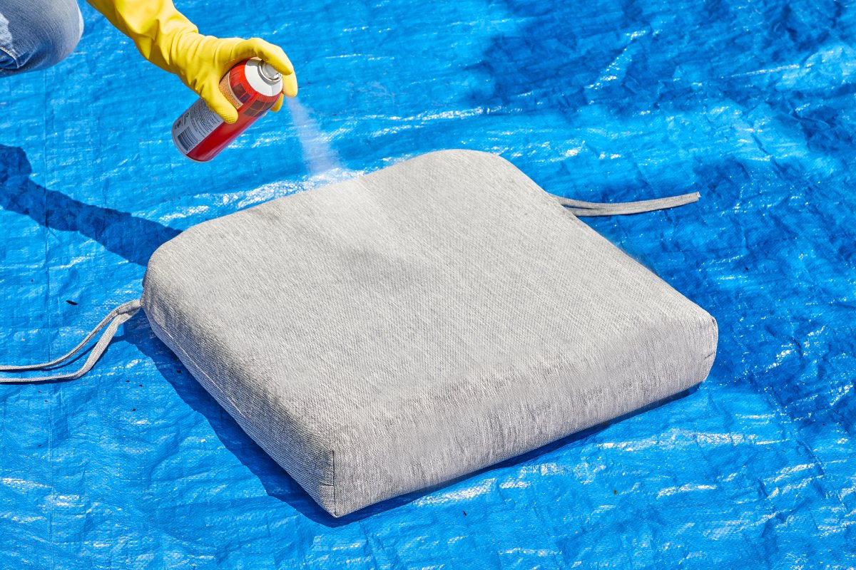 Woman sprays Scotch Guard on a grey patio cushion on a blue tarp.