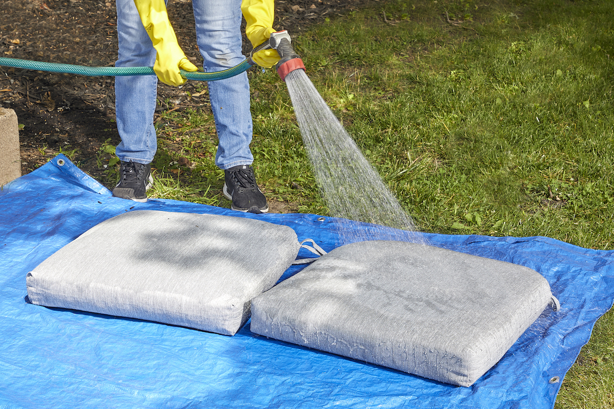 Woman uses garden hose to rinse grey patio cushions outdoors.
