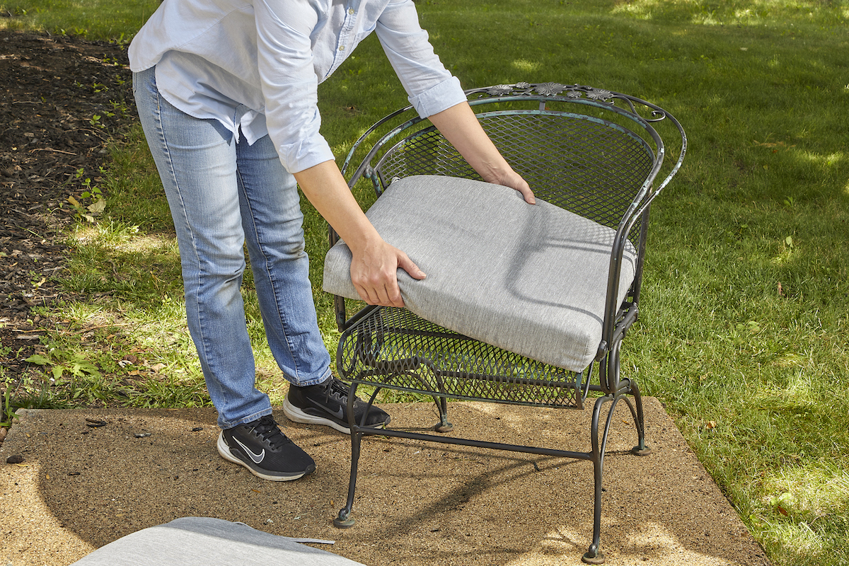 Woman replaces a grey patio cushion on an iron outdoor chair.