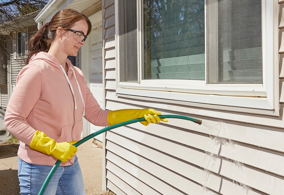 Woman wearing rubber gloves rinses her home's vinyl siding with a garden hose.