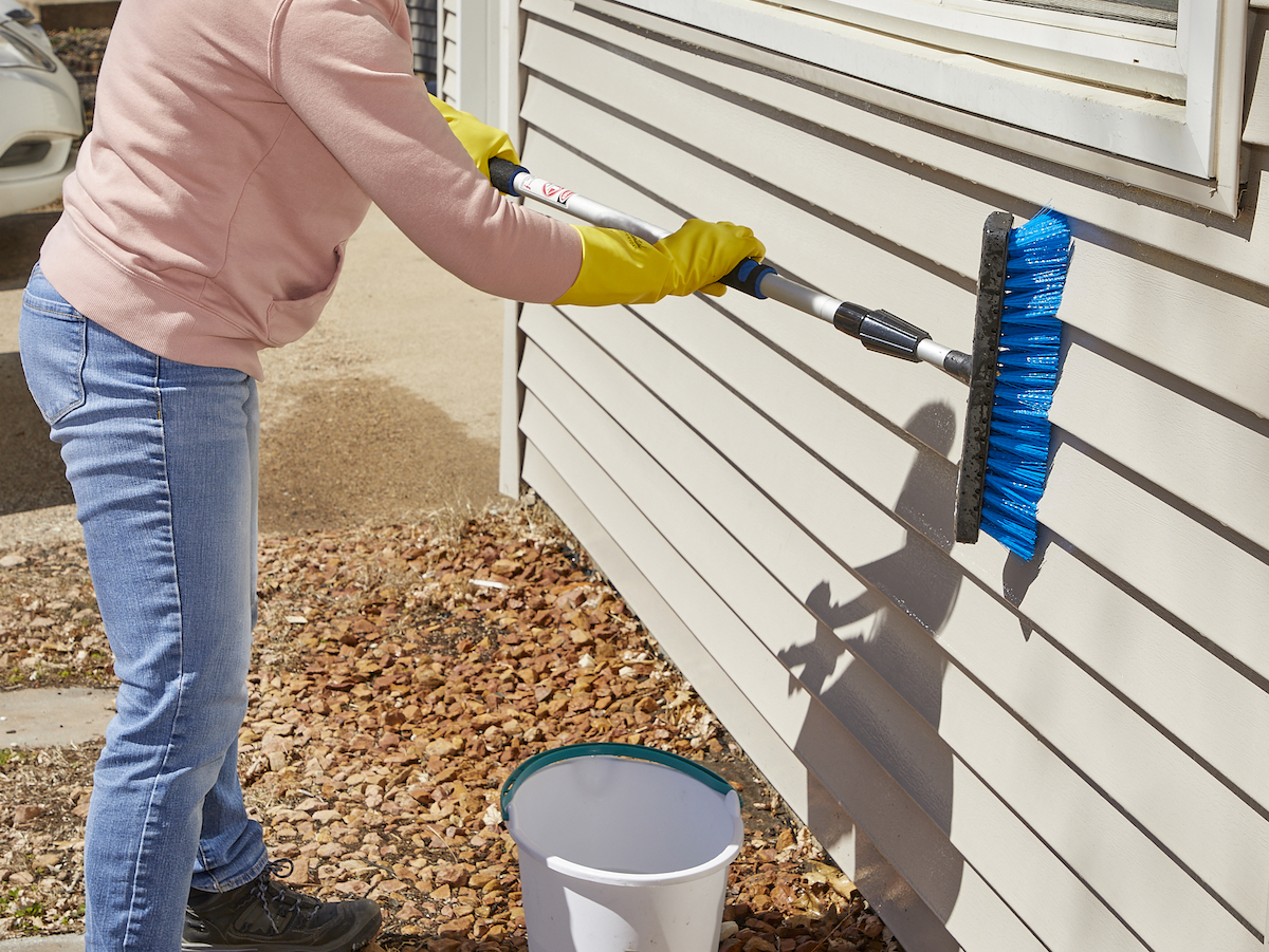 Woman uses soft-bristled brush with long handle to scrub home's vinyl siding.