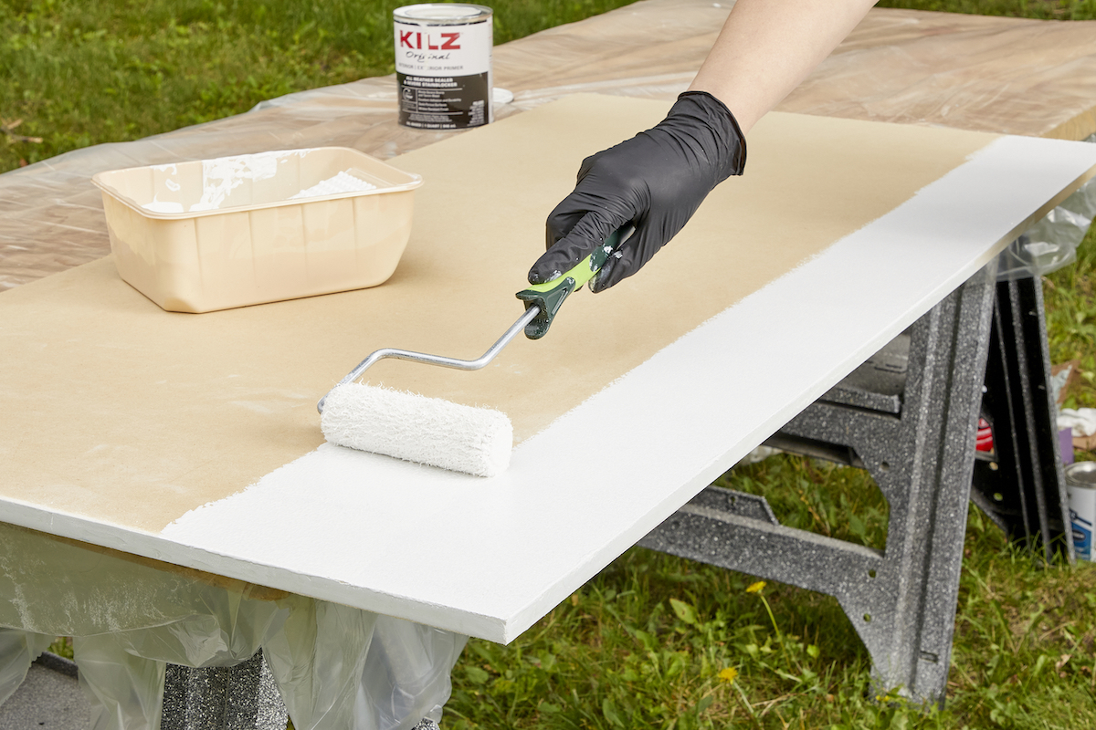 Woman uses a small paint roller to apply primer to a sheet of MDF on a sawhorse.