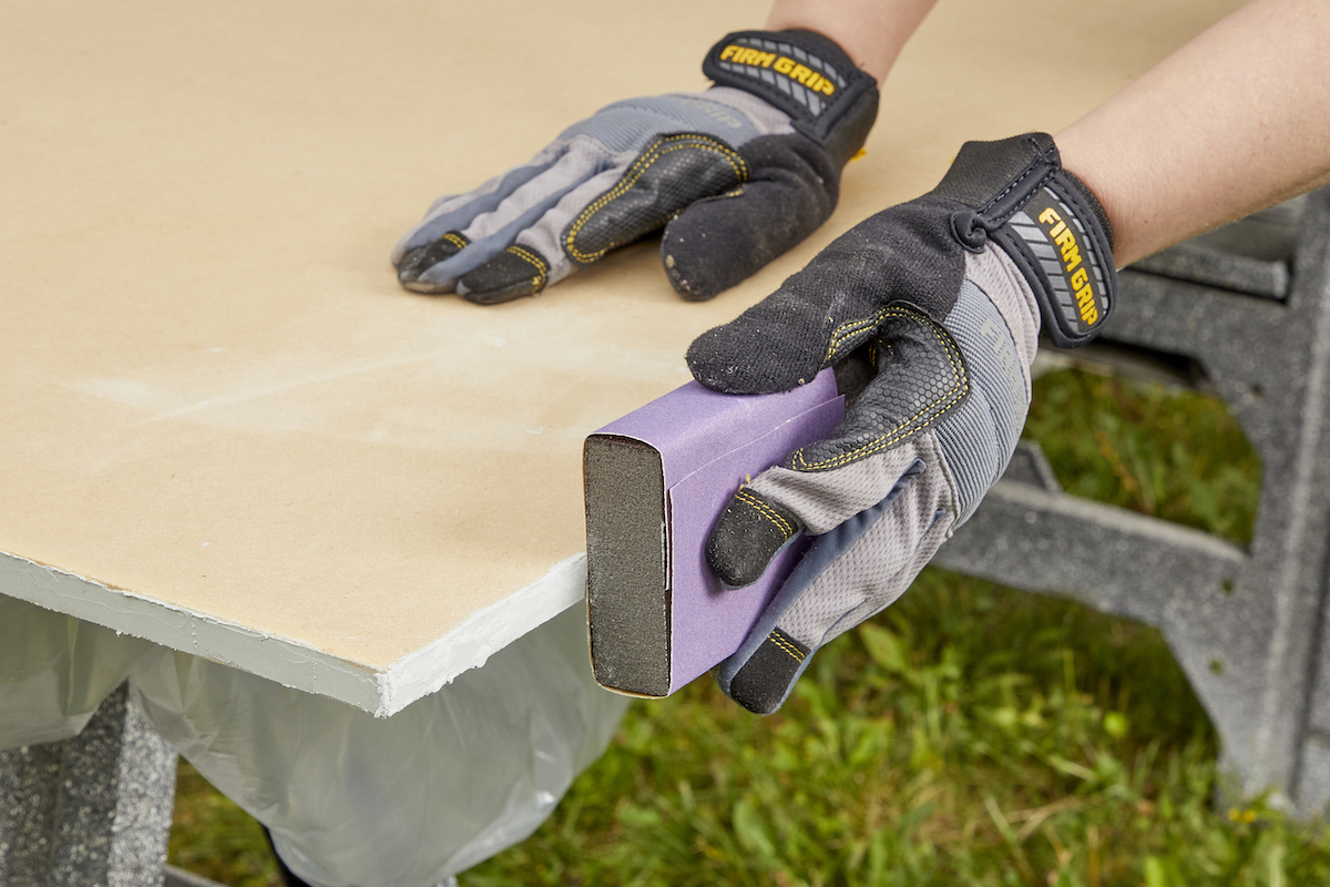 Woman wearing work gloves sands the edges of an MDF board with a sanding block.